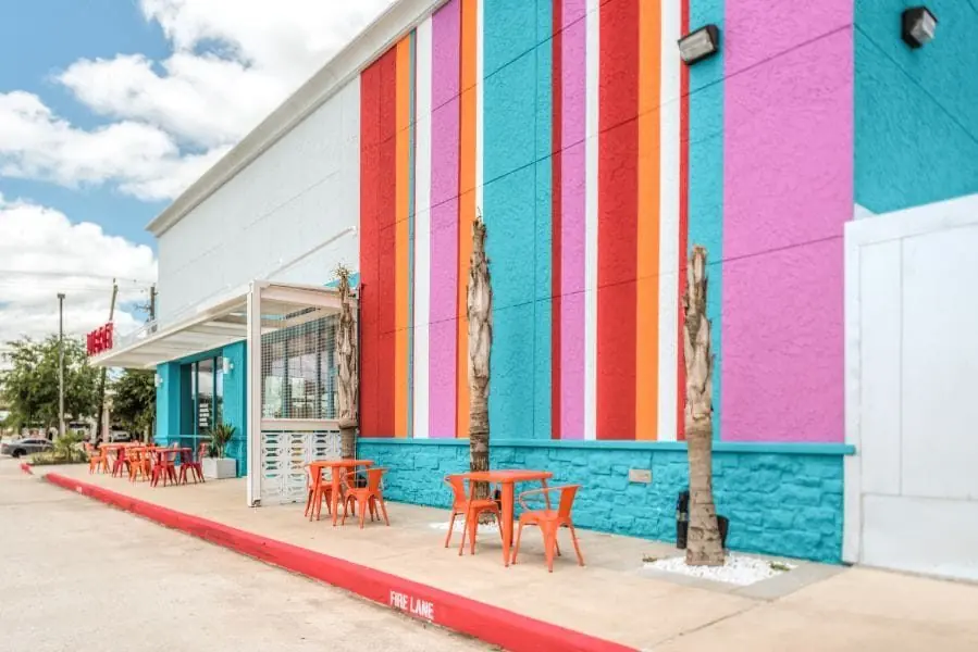 Side facade of Burger Libre in La Porte, TX with vertical color bands of turquoise, orange, magenta, and red, painted block base, and a shaded outdoor patio with orange bistro tables along the sidewalk