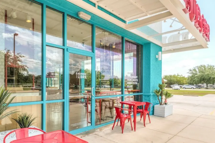 Glass storefront entry of Burger Libre restaurant in La Porte, TX with turquoise aluminum window frames, red Tolix patio chairs, planters with agave, and Lucha Libre branding etched on the glazing