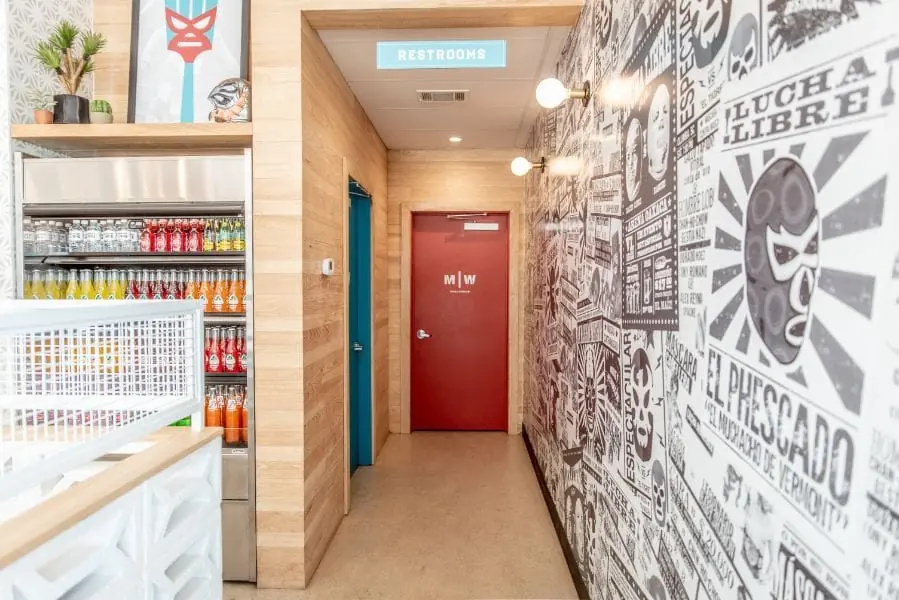 Interior service corridor at Burger Libre in La Porte, TX with a black-and-white Lucha Libre poster mural, refrigerated Mexican soda cooler, wood-paneled walls, and a red restroom door