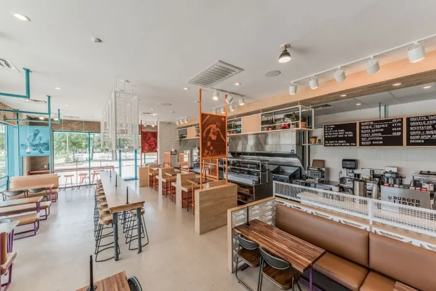Main dining room and counter-service area at Burger Libre in La Porte, TX with chalkboard-style menu boards, an open-concept kitchen, communal wood high-top tables, leather banquette seating, and pendant track lighting