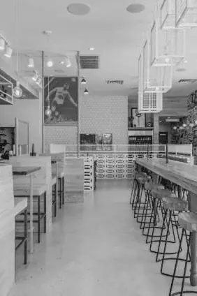 Black-and-white interior view of Burger Libre dining hall in La Porte, TX with long high-top counters, metal bar stools, suspended decorative cube fixtures, and a Lucha Libre wrestler graphic on the back wall