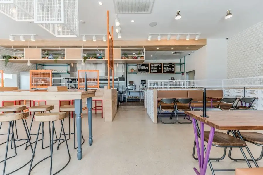 Open dining and ordering area at Burger Libre in La Porte, TX with mixed bar-height and table seating, plant-filled open shelving, decorative breeze-block screens, exposed track lighting, and chalkboard menu boards above the counter