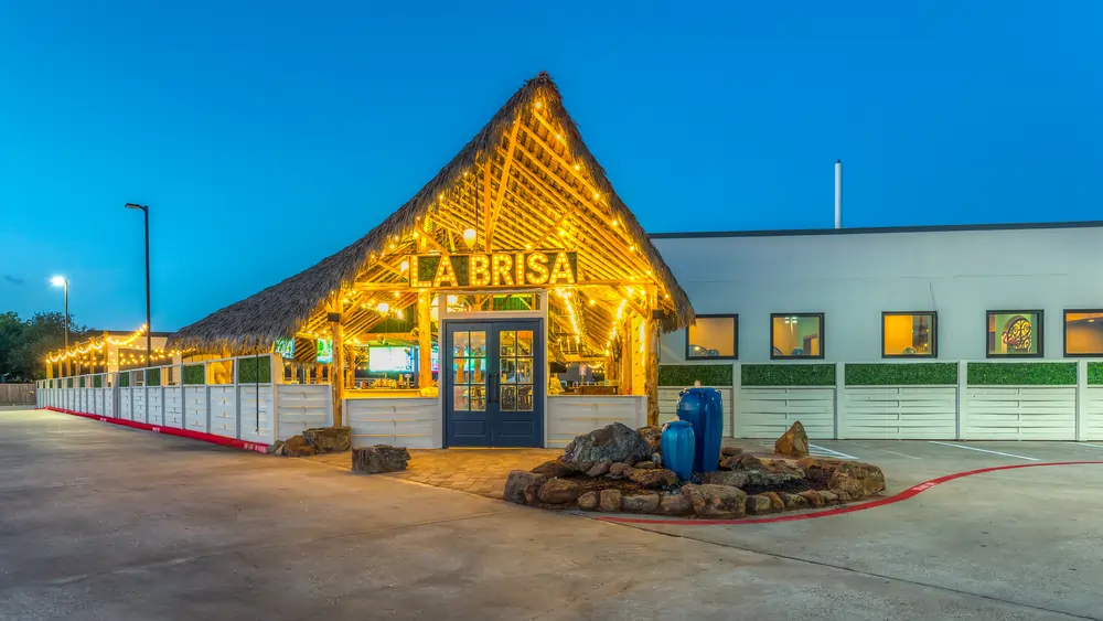 Exterior of LaBrisa Mexican Restaurant in League City, TX at dusk, with an illuminated thatched palapa entry, string lights, and stone water feature