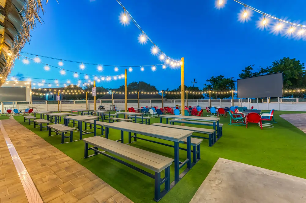 Outdoor yard at LaBrisa with picnic tables on turf, colorful patio chairs, café string lights, and a large outdoor projection screen