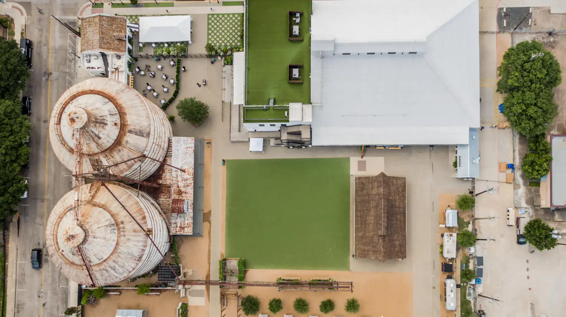 Top-down aerial of Magnolia Market showing the silo roofs, central turf event lawn, thatched pavilion, and rooftop garden terrace on the main warehouse