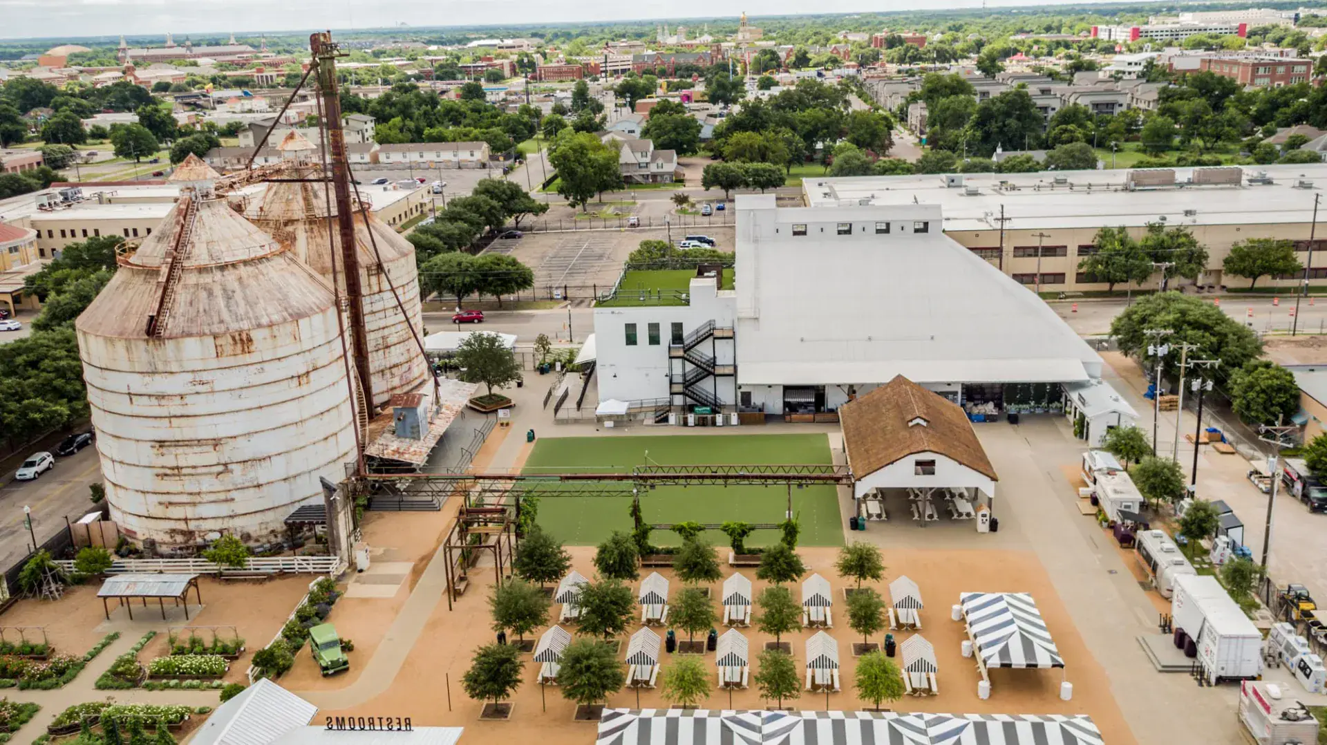 Aerial of the Magnolia Market campus showing the central lawn, striped cabanas, garden beds, and adjacent warehouse with rooftop deck