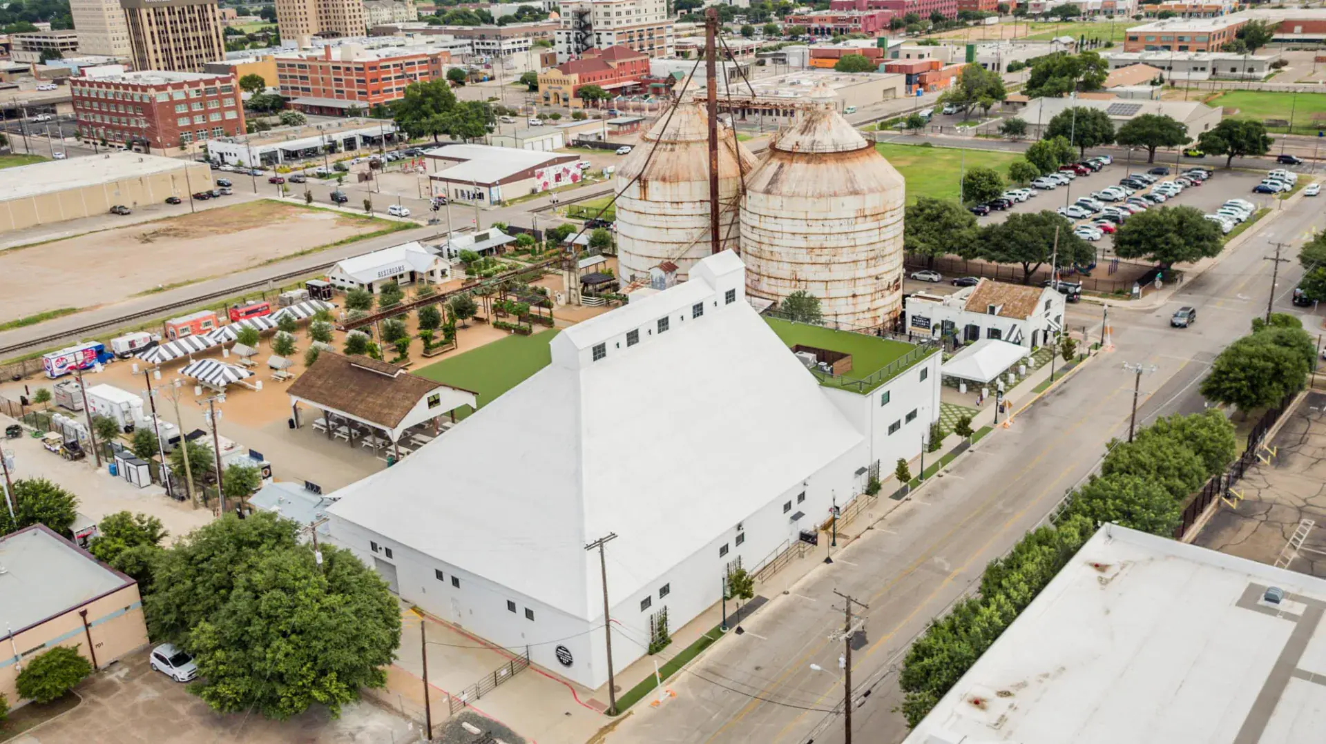 Side-angle aerial of the main Magnolia warehouse with white standing-seam roof, rooftop event deck, and the twin silos and downtown Waco skyline behind