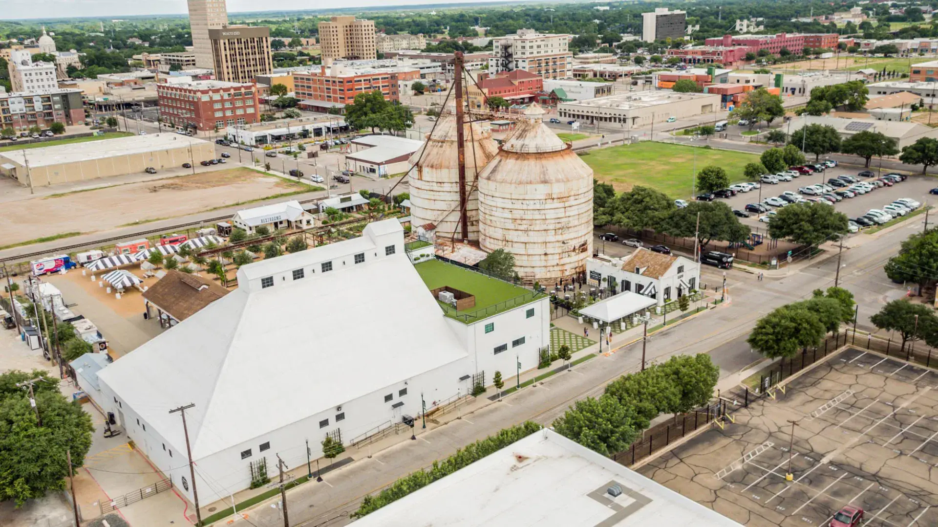 Wider aerial of the Magnolia Market campus with the warehouse, silos, garden, and downtown Waco skyline including the Waco ISD building