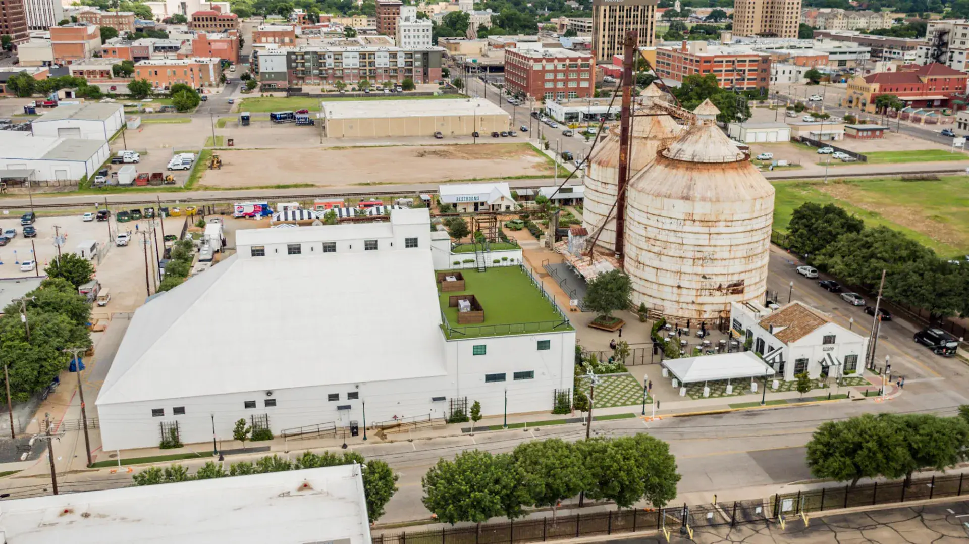 Rear aerial view of Magnolia Market in Waco showing the warehouse footprint, garden beds, food truck park, and railroad alignment along the silos