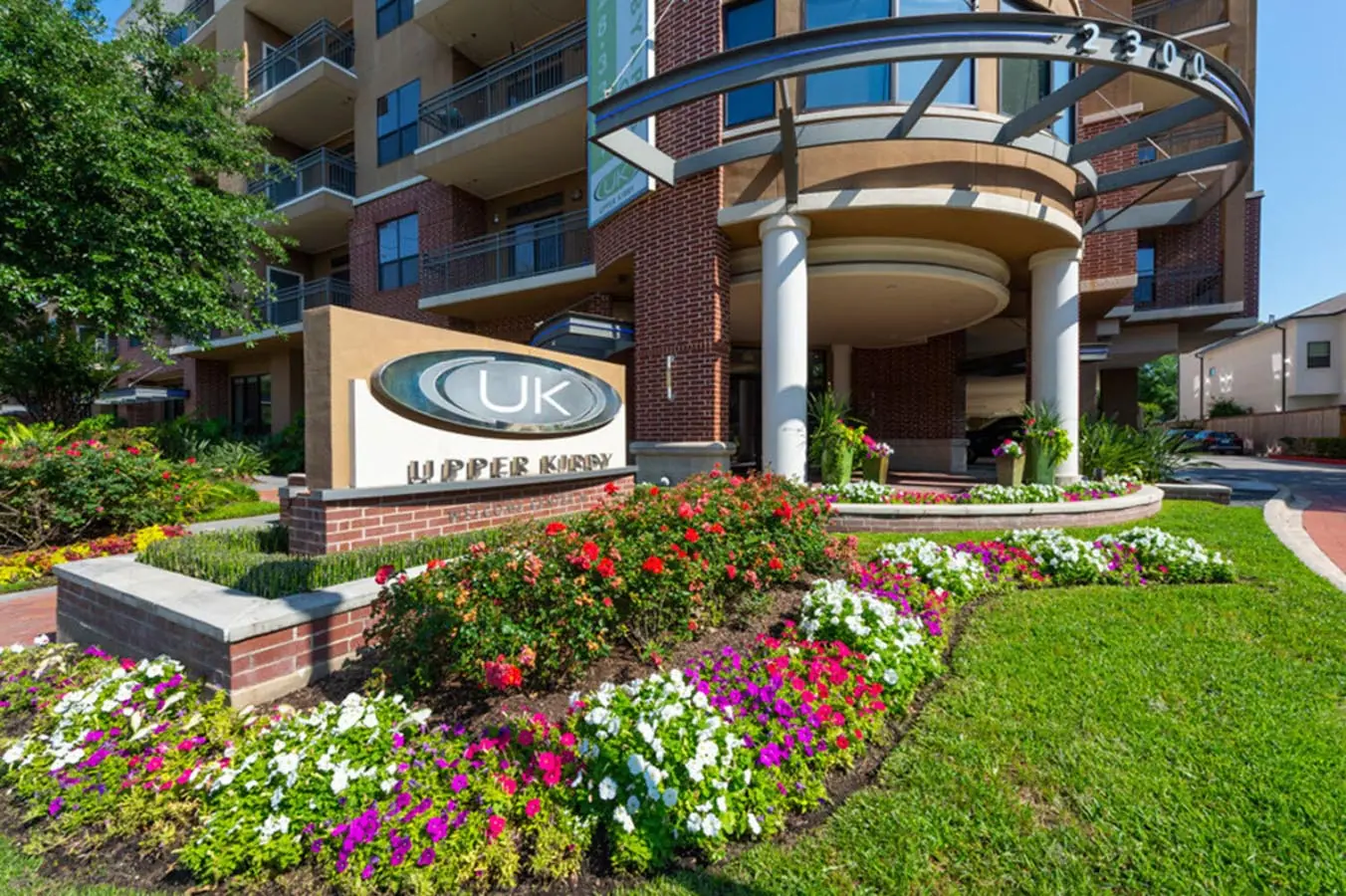Landscaped entry signage at Upper Kirby Apartments in Houston, TX with the UK monogram, flower beds, and the curved glass tower behind