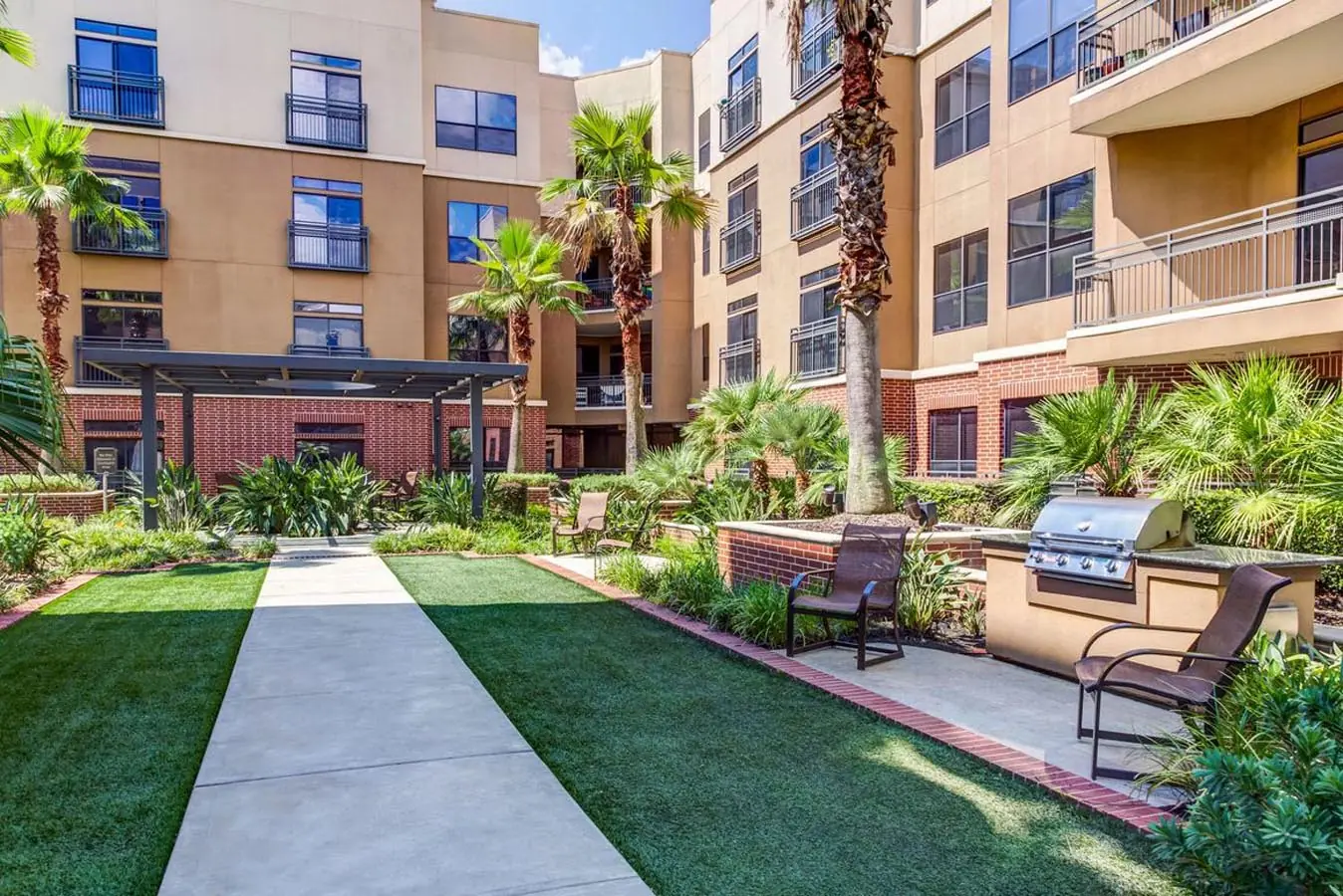 Interior courtyard at Upper Kirby Apartments with a turf lawn walkway, palm trees, brick facade, and built-in grilling stations