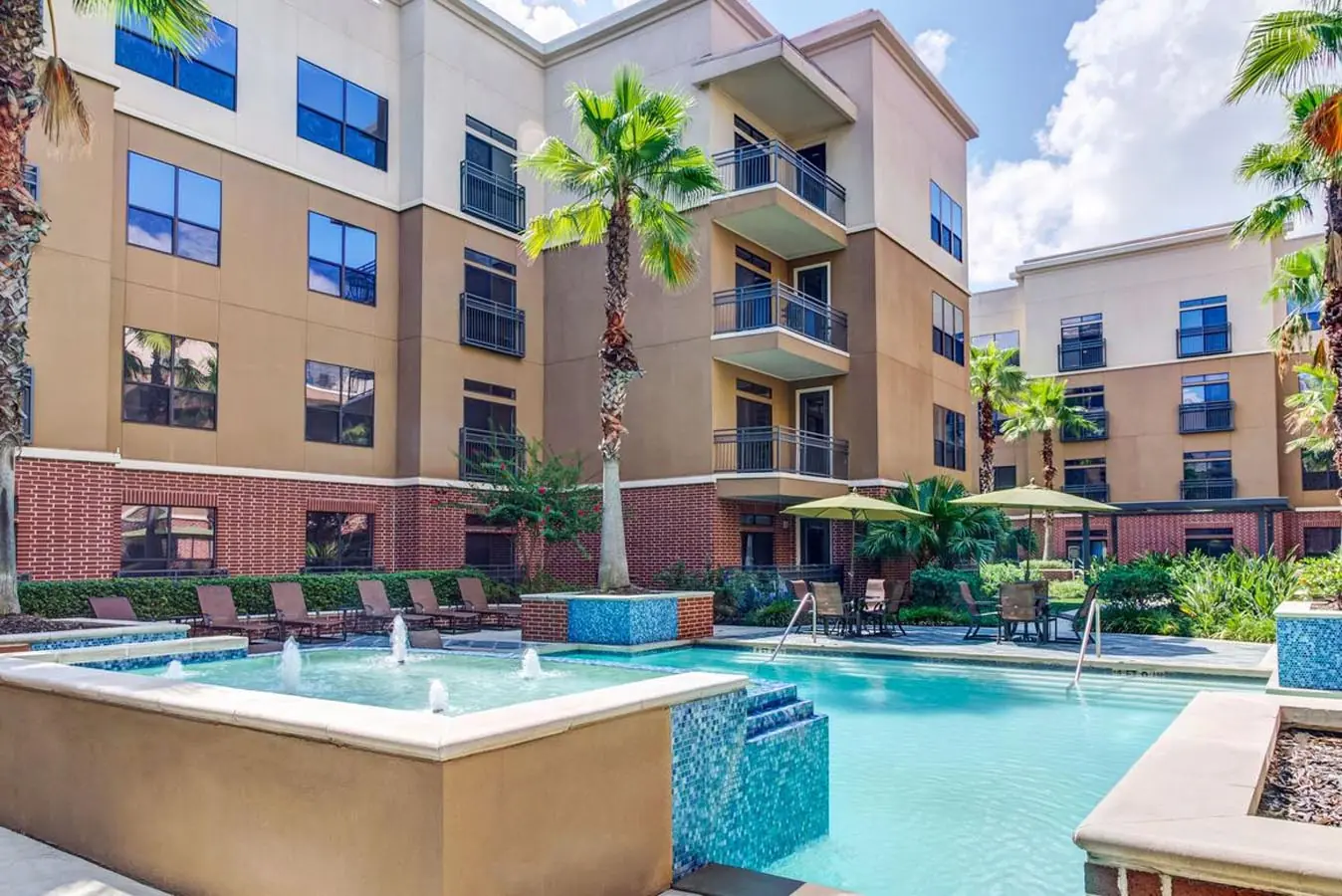 Secondary fountain pool at Upper Kirby Apartments framed by brick and stucco residential balconies, palm trees, and patio umbrellas