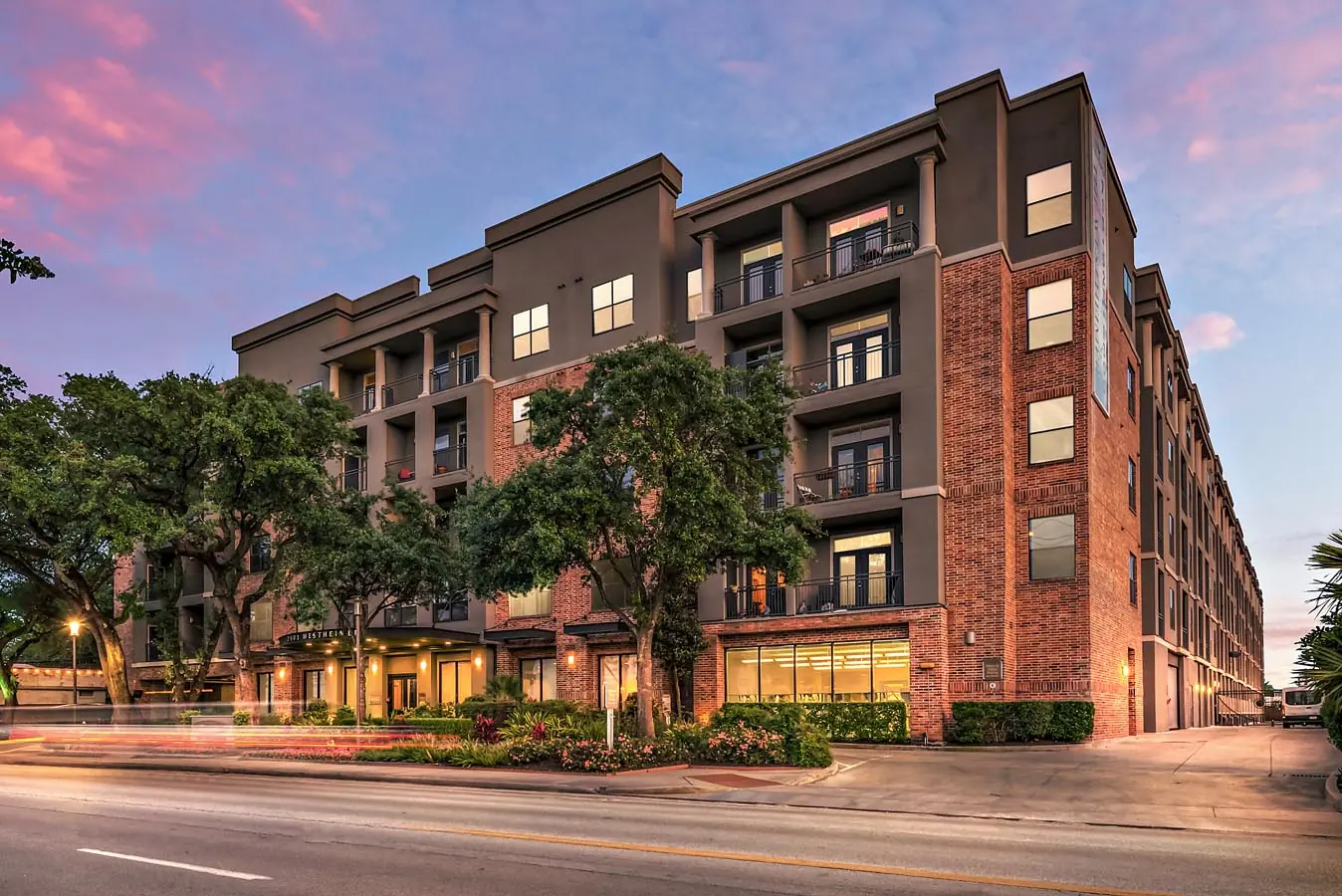 Westheimer Apartments exterior at dusk in Houston, TX with a 4-story wood frame building above a 2-story parking podium and warm street-level lighting